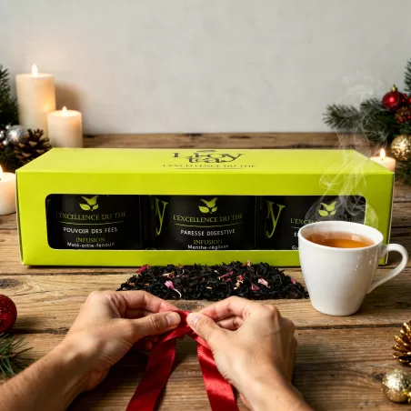 Coffret  L’Excellence du Thé contenant trois boîtes de thé ou infusions, posé au côté d’une tasse fumante et d’un ruban rouge te